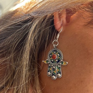 May include: Silver earrings with a hamsa hand design. The hamsa is decorated with blue, green, and yellow enamel and a red stone in the center.
