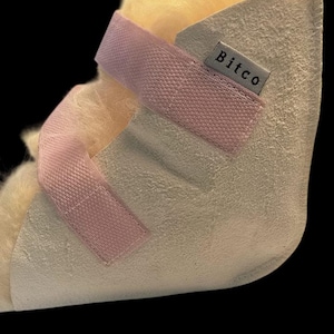 Puede incluir: Una bota médica color crema con una correa rosa y un interior amarillo suave. La etiqueta de la bota dice "Bitco". La bota está diseñada para brindar soporte y protección.