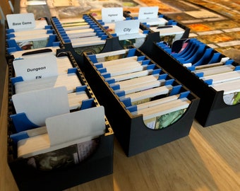 Puede incluir: Cajas de almacenamiento de tarjetas negras con divisores azules y etiquetas blancas. Las etiquetas dicen "Base Game", "Dungeon EN", "City" y "Highland EN".