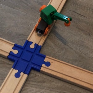 May include: A green toy crane with an orange hook is on a wooden train track. The crane is on a blue and brown track intersection.