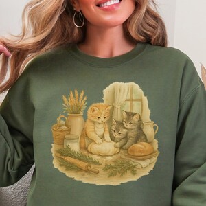 May include: Military green sweatshirt featuring a charming illustration of three kittens gathered around a loaf of bread and a rolling pin. The artwork includes a window, wheat, and a basket. The text "Military Green" is printed on the upper right.