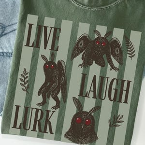 May include: An olive green t-shirt featuring a graphic print of three moth-like creatures with red eyes. The text reads "LIVE LAUGH LURK" in a dark brown serif font. The design includes vertical light green stripes and small fern leaf illustrations.