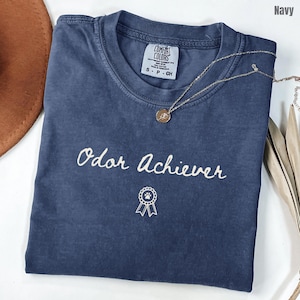 May include: A navy blue short-sleeve t-shirt with a crew neckline and a Comfort Colors brand label. The front features the white text Odor Achiever in a cursive script font, positioned above a small white graphic of a circular award ribbon.