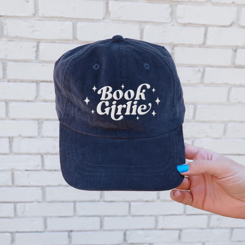 May include: Navy blue corduroy baseball cap with the embroidered text "Book Girlie" in white script. The hat features a curved brim and a small button on top. A stylish accessory for book lovers.