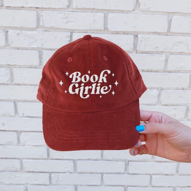 May include: A burgundy corduroy baseball cap with the text "Book Girlie" embroidered in white. The hat has a curved brim and a rounded top. The background is a white brick wall.