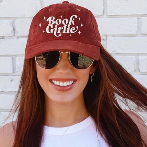 May include: A maroon corduroy baseball cap with the text "Book Girlie" embroidered in white script. The cap features a curved brim and is worn with gold-rimmed sunglasses. The background is a white brick wall.