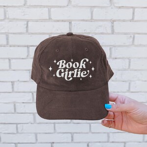 May include: A brown corduroy baseball cap with the text "Book Girlie" embroidered in white. The hat has a curved brim and a button on top. The hat is a casual accessory.