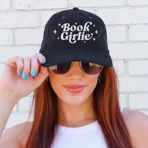 May include: Black baseball cap with the text "Book Girlie" in white script and star accents. The cap is made of a soft material and has a curved brim.