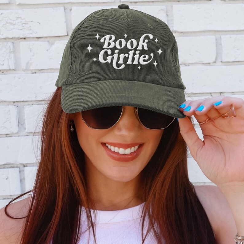 May include: Olive green corduroy baseball cap with the text "Book Girlie" in white script and star accents. The cap has a curved brim and a rounded top. The hat is worn by a person wearing sunglasses.