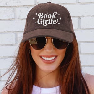 May include: Brown corduroy baseball cap with the text "Book Girlie" in white script. The cap has a curved brim and a button on top. The background is a white brick wall.
