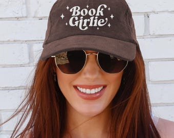 Può includere: Cappellino da baseball in velluto a coste marrone con la scritta "Book Girlie" in bianco. Il cappellino ha una visiera curva e un bottone sulla parte superiore. Lo sfondo è un muro di mattoni bianchi.