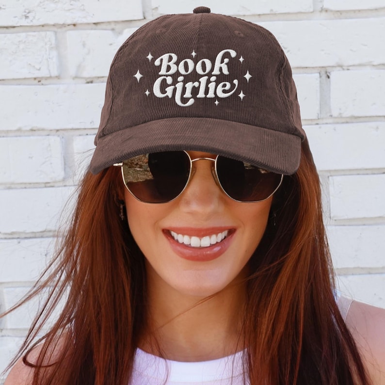 May include: Brown corduroy baseball cap with the text "Book Girlie" in white script. The cap has a curved brim and a button on top. The background is a white brick wall.