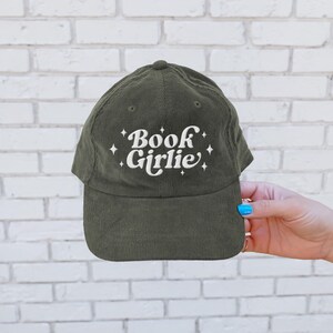 May include: Olive green corduroy baseball cap with the text "Book Girlie" in white script. The text is embellished with small starburst designs. The cap has a curved brim and a button on top.