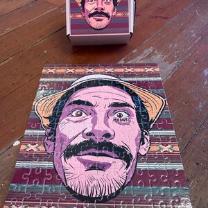 May include: A colorful jigsaw puzzle featuring a cartoon illustration of a person wearing a straw hat and a mustache. The puzzle is set against a background of colorful stripes. The person has the word "MALANDRO" tattooed on their face.