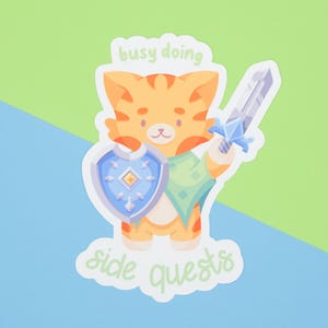 Puede incluir: Una pegatina con un gato atigrado naranja de dibujos animados sosteniendo una espada y un escudo. La pegatina tiene el texto "busy doing side quests" en una fuente caprichosa. El fondo es una división de azul claro y verde.