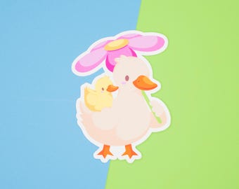 May include: A pink flower with a yellow centre is held above a white duck with an orange beak. A smaller yellow duck with an orange beak is riding on the back of the larger duck.