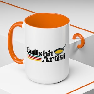 Puede incluir: Taza de cerámica blanca con borde y asa naranja. La taza tiene un gráfico negro que dice "Bullshit Artist" con una sartén estilizada y comida amarilla dentro.