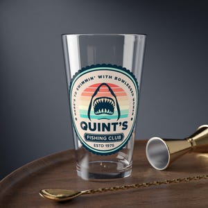 May include: A clear pint glass with a circular graphic featuring a shark's open mouth against a sunset backdrop. The text reads "Quint's Fishing Club ESTD 1975" and "Here's to swimmin' with bowlegged women."