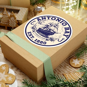 May include: A round sticker with a navy blue and cream design featuring a sailing ship and the words "Antonio Bay Est. 1880 Elizabeth Dane". The sticker is placed on a brown gift box tied with a light green ribbon. Christmas decorations surround the box.