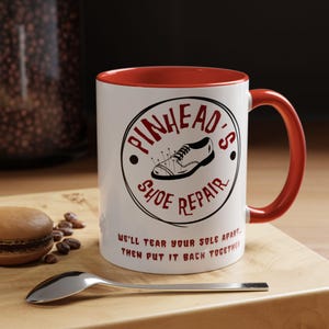 May include: White ceramic coffee mug with a red rim and handle. The mug features a black and white illustration of a shoe with needles in it and the text "Pinkhead's Shoe Repair" in a circle. The text "We'll tear your sole apart, then put it back together" is printed below the circle.