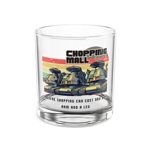May include: A clear glass tumbler with a retro design. The tumbler features a colorful graphic of three futuristic tanks with the words "SHOPPING MALL" and the phrase "WHERE SHOPPING CAN COST YOU AN ARM AND A LEG."