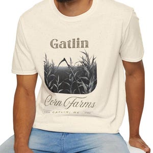 May include: Cream-colored t-shirt featuring a black and white graphic of a cornfield with a scythe and the words "Gatlin Corn Farms." The shirt also includes the text "ESTD GATLIN, NC 1904."