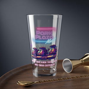 May include: A clear pint glass with a retro design featuring the text "Park Plaza Mall" in pink and blue neon. Below the text is a graphic of three futuristic vehicles. The glass sits on a wooden tray with a gold bar spoon and jigger.