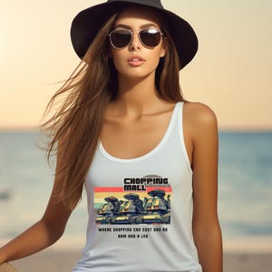 May include: White tank top with a graphic of three robot-like figures and the text "SHOPPING MALL" above the phrase "WHERE SHOPPING CAN COST YOU AN ARM AND A LEG". The tank top is worn by a person wearing sunglasses and a black hat.