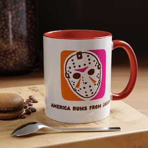 以下が含まれることがあります： 赤い縁と取っ手のある白いセラミック製のマグカップ。マグカップには、ダンキンドーナツのロゴを模倣したホッケーマスクのグラフィックが描かれています。マグカップには「アメリカはジェイソンから逃げる」という文字が印刷されています。