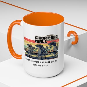 May include: White ceramic mug with an orange interior and handle. The mug features a retro design with the words "SHOPPING MALL" and a graphic of three futuristic tanks. The text below reads "WHERE SHOPPING CAN COST YOU AN ARM AND A LEG."