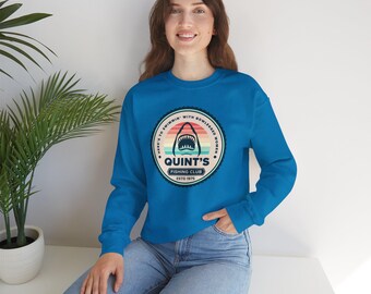 May include: A teal sweatshirt featuring a circular graphic with a shark mouth design and the text "Quint's Fishing Club Estd 1975". The graphic also includes the phrase "Here's to swimmin' with bowlegged women".