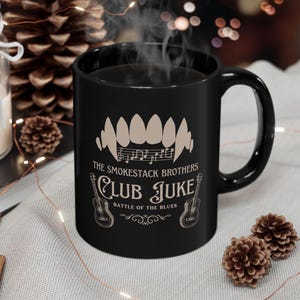 May include: Black ceramic mug with the text "The Smokestack Brothers Club Juke Battle of the Blues" and a graphic of teeth and musical notes. Two guitars are on either side of the text.
