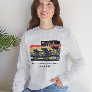 May include: Light gray sweatshirt with a retro graphic that reads "SHOPPING MALL" above a row of robot figures. The text "WHERE SHOPPING CAN COST YOU AN ARM AND A LEG" is below the graphic.