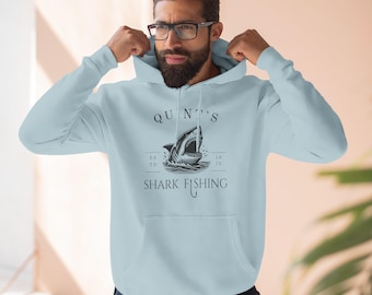 May include: Light blue hoodie with a shark graphic and the text "QUINT'S SHARK FISHING". The graphic features a shark with an open mouth in the ocean. The hoodie has a front pocket and a drawstring hood.