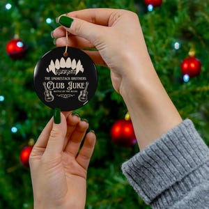 May include: A black round Christmas ornament with gold string. The ornament features white text that reads "The Smokestack Brothers Club Juke Battle of the Blues" with guitar illustrations and a toothy design.