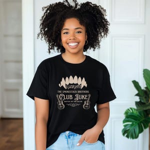 May include: Black t-shirt with the text "The Smokestack Brothers Club Juke Battle of the Blues" in a vintage style. The design includes a stylized mouth, musical notes, and two guitars. The shirt is worn by a person with curly hair.