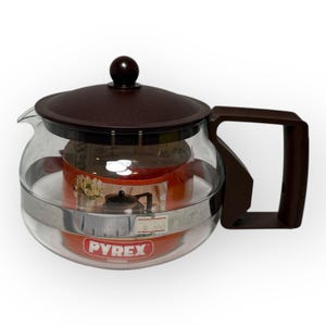 Può includere: Teiera Pyrex in vetro trasparente con coperchio e manico marroni. La teiera ha una forma rotonda e una fascia metallica al centro. All'interno c'è un contenitore rosso con un disegno floreale. Il logo Pyrex è visibile.
