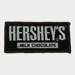 May include: A rectangular embroidered patch with a black background and silver-white stitching. The text HERSHEY'S is prominently displayed in large capital letters, with the words MILK CHOCOLATE printed in a smaller font on a rectangular bar below.