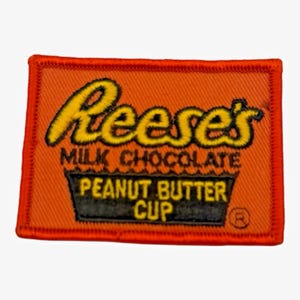 May include: A rectangular embroidered patch with an orange background. The text Reese's is stitched in yellow with a black outline. Below it, the text MILK CHOCOLATE PEANUT BUTTER CUP is stitched in yellow inside a black trapezoid shape.