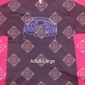 May include: A dark gray t-shirt with a V-neck and a graphic design in light purple. The design features the words "DUNK DOPE3" within the outline of goggles. The shirt is labeled "Adult Large".