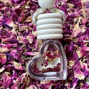May include: A clear glass heart-shaped bottle filled with dried rose petals, hanging from a white wooden bead and a string with gold accents. The bottle is surrounded by a bed of dried rose petals.