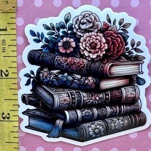May include: A stack of five vintage books with ornate covers, adorned with a bouquet of pink, red, and blue flowers. The books are arranged in a pyramid shape, with the flowers cascading over the top.