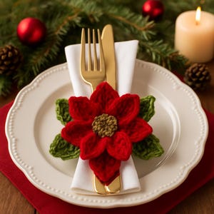 May include: A Christmas table setting with a white plate, gold cutlery, and a white napkin. A red crocheted poinsettia flower with green leaves and a brown center adorns the napkin. The plate sits on a red placemat.