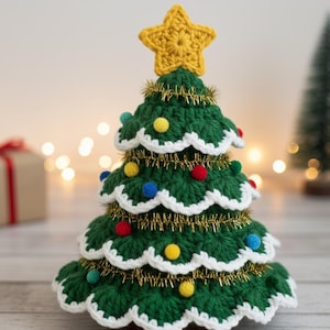 Könnte beinhalten: Ein handgefertigter gehäkelter Weihnachtsbaum mit einem gelben Sternaufsatz und bunten Pompon-Ornamenten. Der grüne Baum ist mit goldenem Lametta und weißen, gewellten Kanten verziert und schafft eine festliche Weihnachtsdekoration.