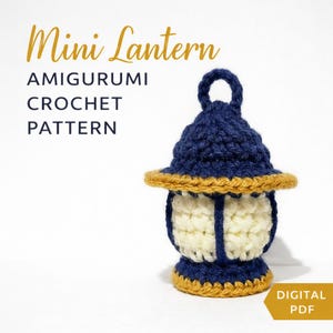Puede incluir: Una mini linterna de ganchillo en azul marino, crema y dorado. La linterna tiene una parte superior redondeada con un lazo, un centro color crema y un ribete dorado. El texto "Mini Lantern Amigurumi Crochet Pattern" es dorado.