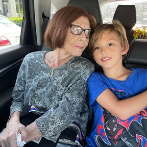 May include: A woman with short brown hair and glasses is sitting in the back seat of a car with a young boy. The woman is wearing a grey floral shirt and the boy is wearing a blue shirt with a Spiderman graphic.