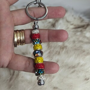 May include: A silver keychain with a beaded charm. The charm is made of colorful beads with rhinestones. The beads are red, yellow, blue, and green.