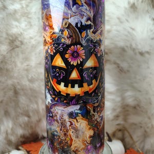 May include: A cylindrical tumbler featuring a glossy finish with a swirling marble pattern in shades of purple, orange, and black. The center displays a black jack-o'-lantern graphic adorned with colorful floral accents and glitter.