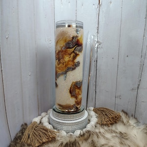 May include: A tall, clear tumbler with a lid and straw. The tumbler features a swirling design of brown, tan, and white, with glitter accents. It sits on a mirrored base and a decorative beaded ring.