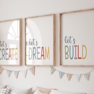 May include: Three framed art prints with the phrases "let's CREATE", "let's DREAM", and "let's BUILD" in colourful letters. The prints are in light wood frames and are hung on a white wall. A banner of small flags hangs below the prints.